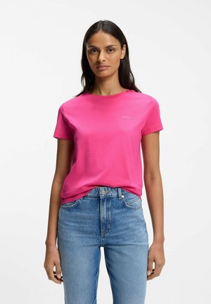 Woman wearing bright pink BOSS short-sleeve t-shirt tucked into high-waisted blue jeans, standing against plain white background.