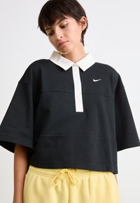 Čierne skrátené polo tričko s bielym golierom a predným zapínaním; na ľavej hrudi obsahuje malé logo Nike. Mäkko zamatový pocit, voľný strih.