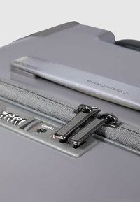 Grauer Koffer mit glatter Oberfläche, einem Reißverschluss aus Stoff, einem Zahlenschloss und schwarzen Reißverschlüssen mit dem eingeprägten Markennamen "Piquadro".