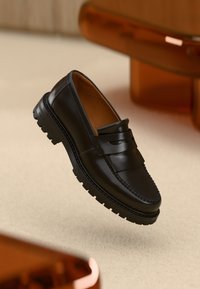 Black leather loafers with a decorative strap, textured sole, and distinct stitching detail. Floating against a neutral backdrop.