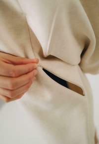 Krémszínű kapucnis pulóver, oldalán cipzáras zsebbel. Az anyag lágy és texturált. Egy kéz húzza a cipzárt, felfedve a fekete bélést belül.