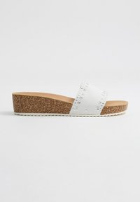 Sandalia deslizante blanca con una correa texturizada que presenta tachuelas plateadas, suela de corcho en cuña y forro interior suave. Tacón plano y forma redondeada.