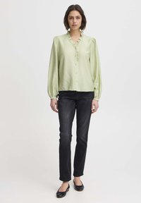 Light green, long-sleeve blouse with a ruffled V-neck and button front, paired with black slim-fit jeans and black flat shoes.