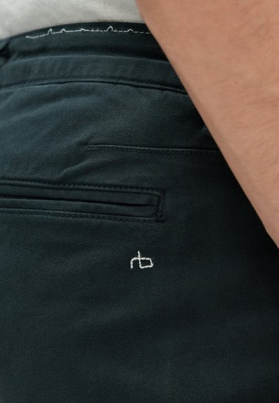 Tamsiai žali kelnių su lygiu paviršiumi, turinčių užpakalinį kišenę ir mažą baltą siuvinėtą logotipą šalia kišenės siūlės.