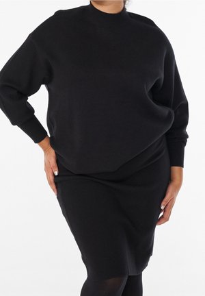 Femme portant un pull noir à manches longues et une jupe assortie à hauteur des genoux, debout avec une main sur la hanche sur fond blanc.