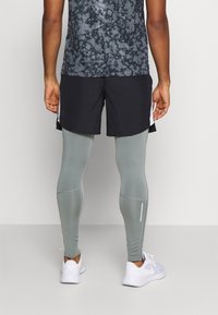 Tenue de sport comprenant un haut gris à motifs, un short noir et un legging vert clair avec une bande réfléchissante, associé à des baskets blanches.