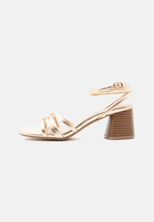 Kultainen remmikenkä, jossa on paksu puinen korko ja nilkkaremmi soljella, yksinkertaisella vaalealla taustalla.