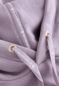 Lyse lilla sweatshirtstof med matchende snore trukket gennem guldfarvede metaløjer, trykt med "B Y TED BAKER" i hvidt.