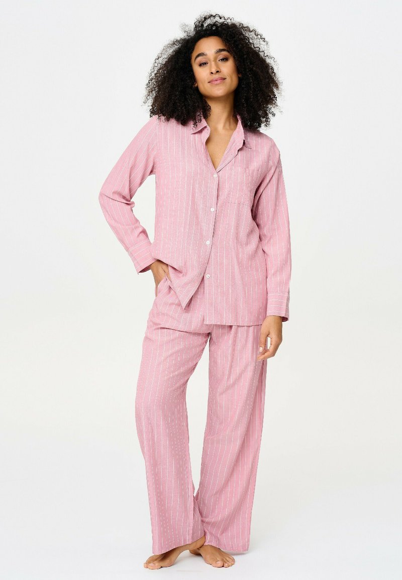 Femme debout pieds nus portant une chemise à manches longues rayée rose à boutons et un pantalon ample assorti sur un fond blanc uni.