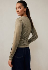 Gari piedurkņu krekls olīvu zaļā krāsā ar piegulošu dizainu un savilktām detaļām, kopā ar tumši zilām, platu kāju džinsu biksēm. Gluda tekstūra.