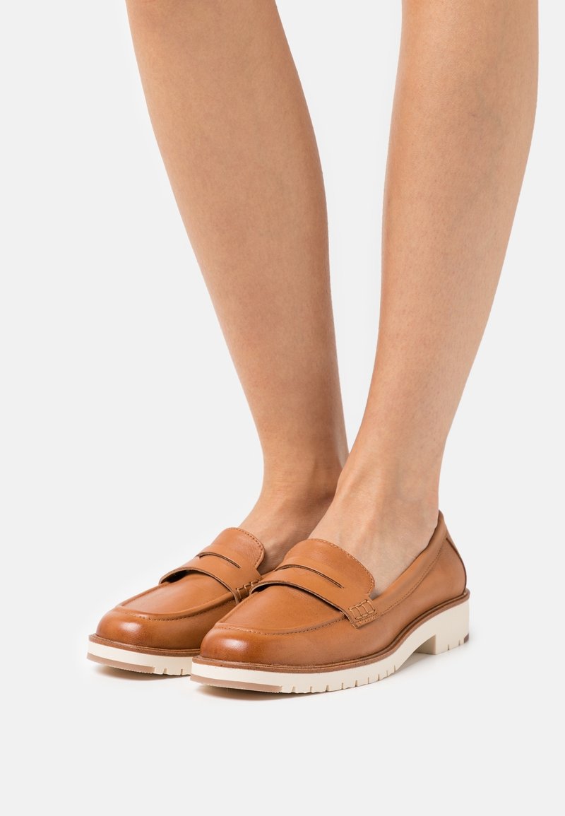 Mocassins en cuir marron avec une finition texturée, présentant un design cousu et une semelle en caoutchouc blanche. Silhouette minimaliste avec des découpes sur les côtés.