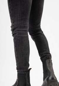 Jean skinny en denim noir avec un motif déchiré sur la cuisse gauche, associés à des bottines noires à talon bas avec des côtés élastiques et des semelles texturées.