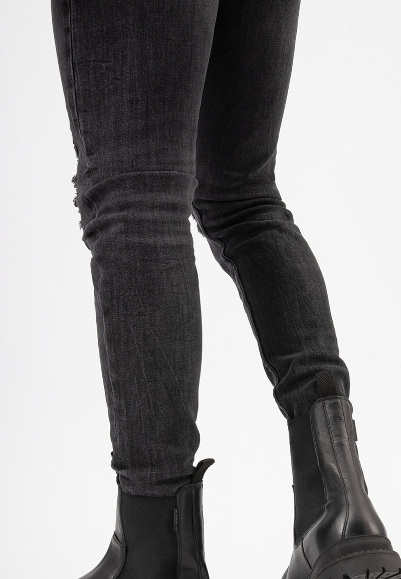 Jean skinny en denim noir avec un motif déchiré sur la cuisse gauche, associés à des bottines noires à talon bas avec des côtés élastiques et des semelles texturées.