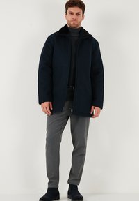 Námornícka fleece bunda s čiernym golierom, so zipsovým predným zapínaním a dvoma bočnými vreckami, v kombinácii so šedými zúženými nohavicami a čiernou obuvou.