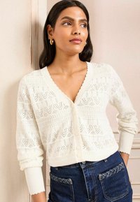 White knitted cardigan with a V-neck, scalloped edges, and textured pattern. Features buttons and long sleeves, paired with dark denim jeans.
