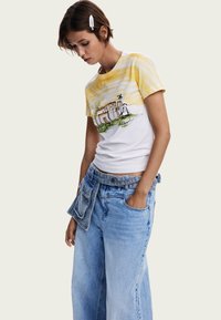 Vit bomullströja med gula randiga ärmar, med ett tryckt arkitektoniskt mönster. Matchad med lösa blå denimjeans och ett denimbälte.