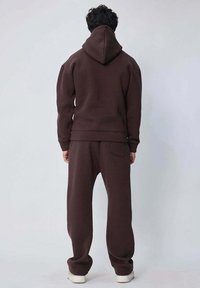 Haut à capuche marron avec une grande capuche, un pantalon de survêtement marron assorti et des baskets blanches. Tissu doux, coupe décontractée, poche arrière visible sur le pantalon.