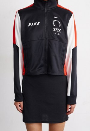 Femme portant une veste zippée Nike noire, blanche et rouge avec logos, associée à une jupe noire, debout devant un fond uni.
