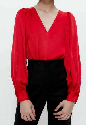 Top in raso rosso con scollatura a V, maniche lunghe plissettate e vita aderente, abbinato a pantaloni neri sartoriali. Texture liscia, design elegante.