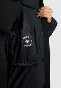 Lange zwarte jas met een gladde buitenkant en een glanzende binnenkant. Beschikt over een wit logo met een blad en de tekst "KHUJO ORIGINAL."