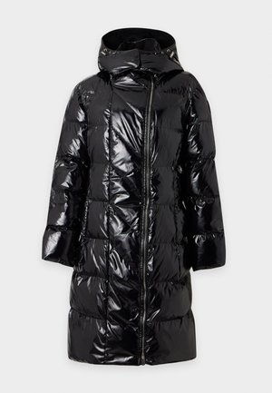 Femme portant un long manteau matelassé noir, une chemise rose, un pantalon noir avec ceinture et des bottes noires, debout devant un fond clair uni.