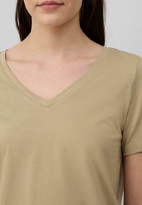 Beige V-Ausschnitt-T-Shirt aus weichem Stoff mit kurzen Ärmeln, das eine glatte Textur und subtile Nähte entlang des Ausschnitts aufweist.