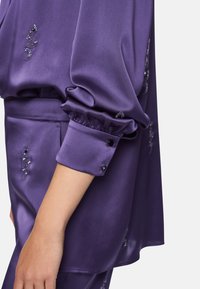 Purple satin blouse with gathered cuffs. Features decorative embellishments and a smooth texture. Subtle shine enhances design details.