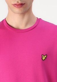 Fucsia színű pamut póló kerek nyakkivágással, bal oldalon egy kis fekete és sárga hímzett saslogóval. Sima textúra.