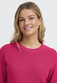 Vruće ružičasti pamučni džemper s okruglim izrezom i teksturiranom tkaninom. Žena s malim zlatnim naušnicama u obliku obruča, valovite kose i prirodnim make-upom.