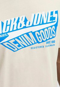 Krémové tričko s modrým a bílým grafickým textem. Zobrazuje "CK&JONES" a "DENIM GOODS" tučným, dynamickým písmem, hladká bavlněná textura.