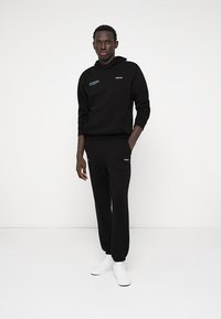 Zwart hoodie en sportbroek set, gemaakt van katoen, met een voorzak, geborduurde logo's in blauw en wit, gecombineerd met witte sneakers.