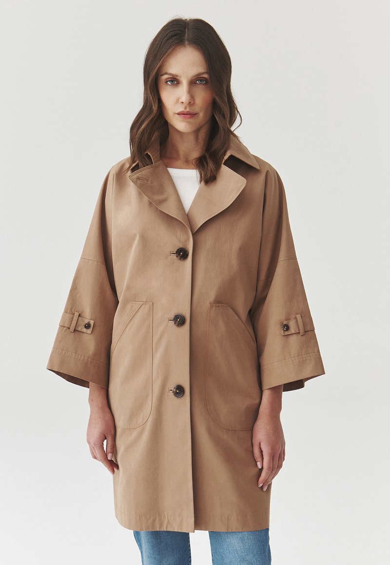 Manteau oversize en tissu lisse de couleur beige, avec un large col, quatre boutons, de grandes poches et des manches courtes agrémentées de sangles.