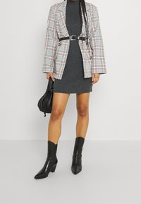 Blazer à carreaux avec boutons en or, robe grise à côtes, ceinture noire, et bottes noires à talons pointus avec une finition texturée. Sac à main noir.