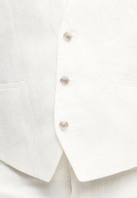 Κοντινή λήψη λευκού γιλέκου με τρία διαφανή κουμπιά και ορατές τσέπες μπροστά, που δείχνει την υφή του υφάσματος και τις άκρες του ρούχου.