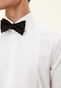 Weißes Hemd mit vertikalen Falten, ausgestattet mit einer schwarzen Satin-Schleife. Button-Down-Kragen und glatte Stofftextur.