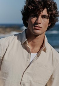 Chemise beige clair boutonnée avec une poche poitrine, portée sur un débardeur blanc. Tissu doux, cadre naturel avec un arrière-plan flou d'océan.