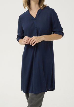 Kobieta ubrana w granatową tunikę do kolan z podwiniętymi rękawami i szare spodnie na białym tle.