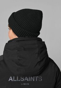 Svart stickad mössa med ribbad textur. Personen bär en svart huvtröja med "ALLSAINTS LONDON" logotyp på ryggen.