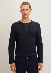 Camisa henley de manga larga en color azul marino con tres botones, que cuenta con un cuello redondo y rayas sutiles en los hombros, combinada con pantalones oscuros.