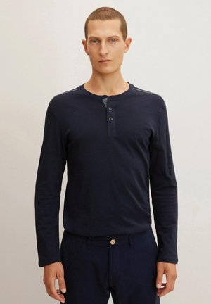 Camisa henley de manga larga en color azul marino con tres botones, que cuenta con un cuello redondo y rayas sutiles en los hombros, combinada con pantalones oscuros.