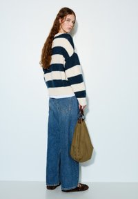 Pruhovaný pletený svetr v námořnické modré a krémové barvě, doplněný širokými džínami s volnými nohavicemi. Modelka drží olivově zelenou tašku s kovovými detaily.