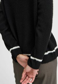 Maglione nero lavorato a maglia con strisce bianche ai polsini e all'orlo. Tessuto testurizzato, vestibilità sopraffina, maniche lunghe, indossato con le mani incrociate dietro.