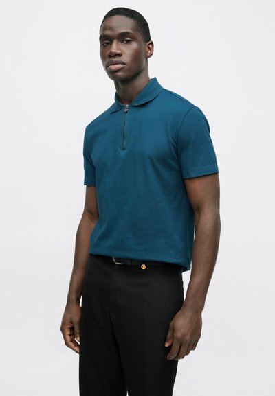 Mėlynos žalsvos spalvos polo marškinėliai su užtraukiamu apykakle, trumpomis rankovėmis ir subtiliu prekės ženklo siuvinėjimu. Dėvimi su juodomis kelnėmis ir diržu su aukso spalvos akcentu.