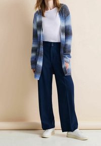 Knälång kofta i gradientblått stickat, i kombination med vida marinblå byxor och vita sneakers. Släta texturer och avslappnad passform synliga.