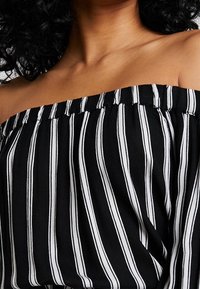 Top asymétrique en tissu rayé noir et blanc, avec un col froncé et une texture douce. Accent sur les rayures verticales.