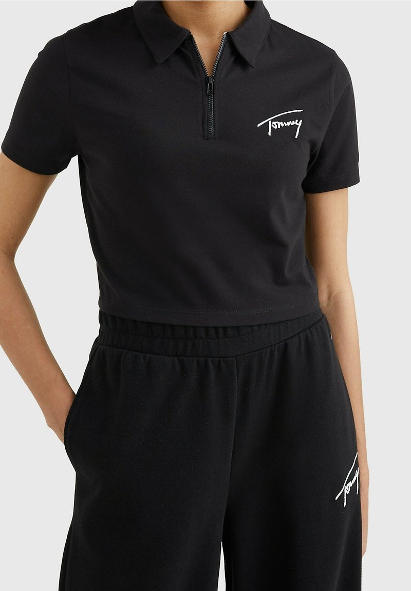 Černé zkrácené polo tričko se zipovým límcem, s bílou výšivkou loga "Tommy". Nošeno s odpovídajícími černými tepláky. Měkký bavlněný materiál.