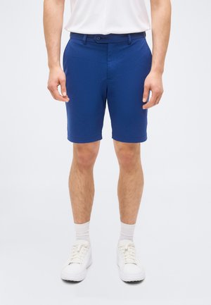 Bărbat purtând pantaloni scurți albaștri croiți pe măsură, șosete albe, adidași albi și o cămașă albă băgată în pantaloni, stând în fața unui fundal alb.