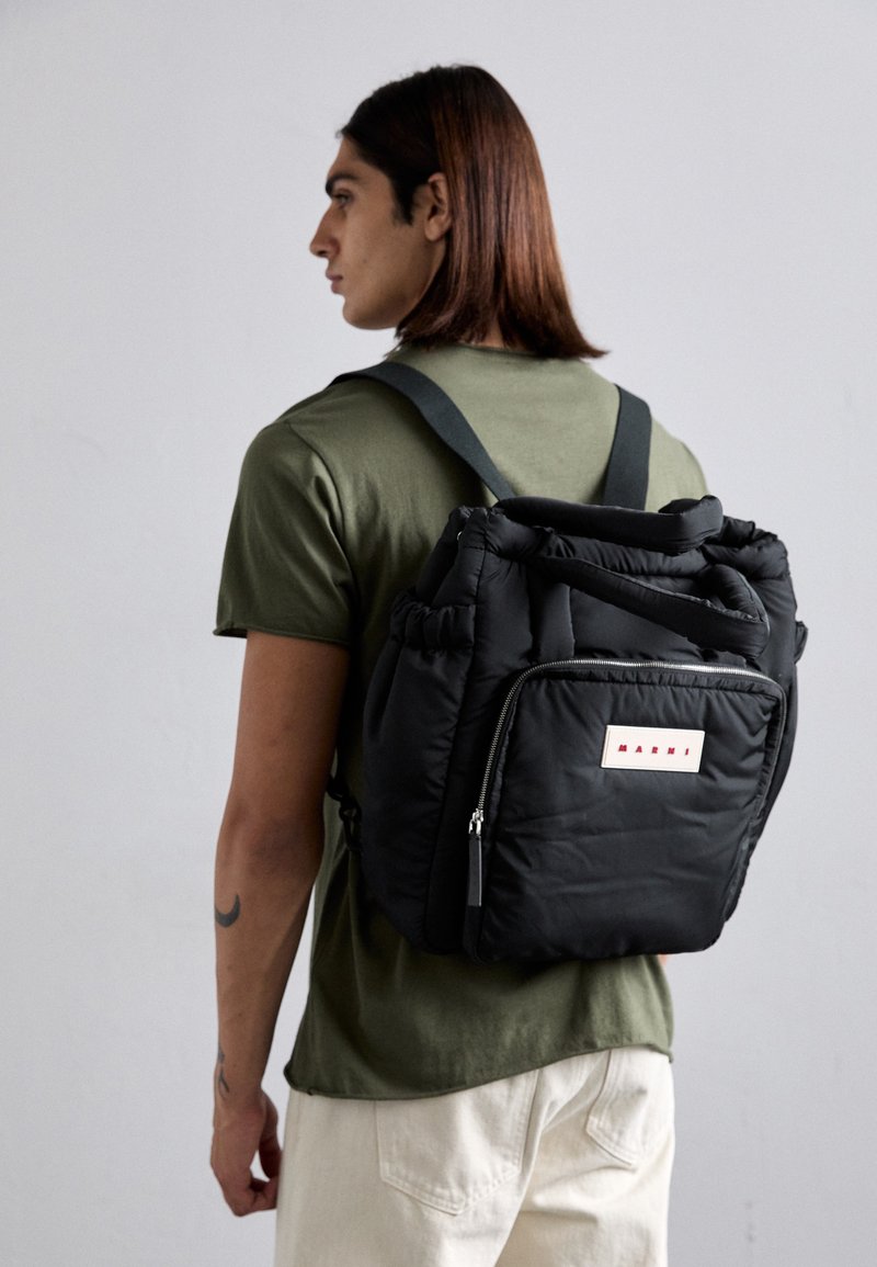 Black padded backpack with adjustable straps, front zipper pocket, and logo tag. Worn on the back of a person in an olive green shirt.