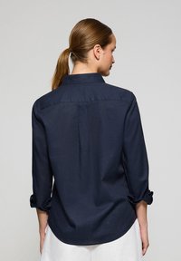 Navy blue shirt made of lightweight fabric, featuring a collared neckline, long sleeves rolled up, and a rounded hem at the back.