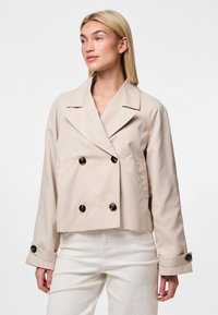 Donna che indossa un giubbotto beige doppiopetto con bottoni neri e pantaloni bianchi, che guarda leggermente a sinistra su uno sfondo semplice.